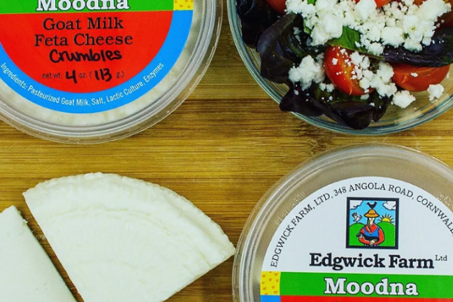 Two containers of goat milk feta cheese and a salad with feta on a wooden surface.
