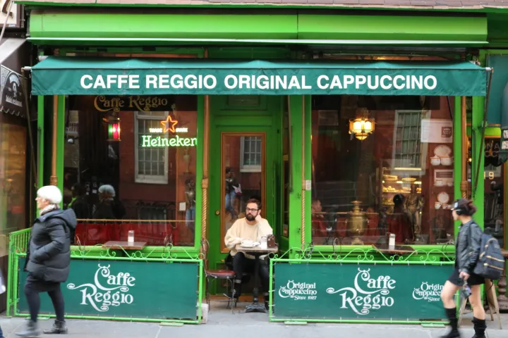 Street view of Caffe Reggio, a man seated outside with green decor and pedestrians passing by.