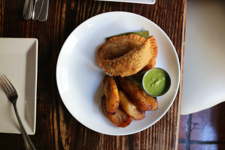 Empanadas and fried plantains on a white plate with green sauce.