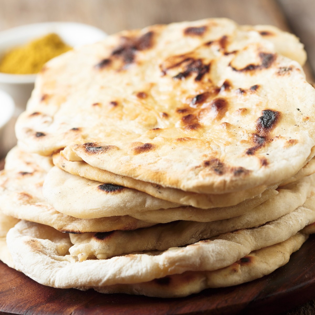 Stack of naan bread on a wooden board with small bowls of spices in the background.
