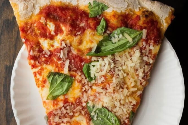 Slice of pizza with basil and cheese on a paper plate, wooden table background.