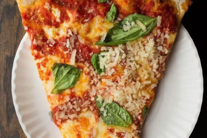 Slice of pizza with basil and cheese on a paper plate, wooden table background.