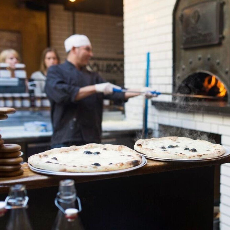Chef baking pizzas in a wood-fired oven with pizzas on the counter.
