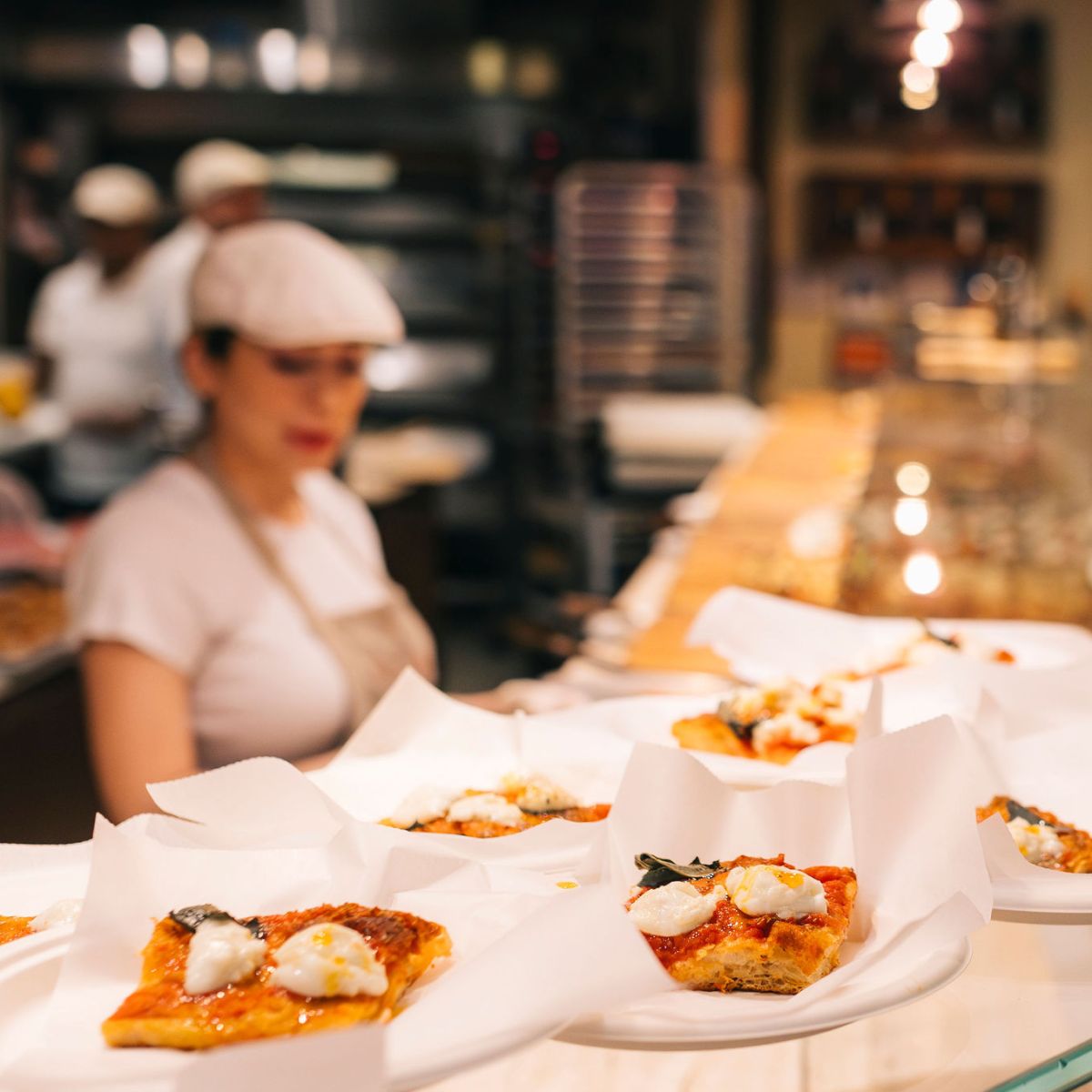 Pizza slices on plates in a restaurant with chef in background.