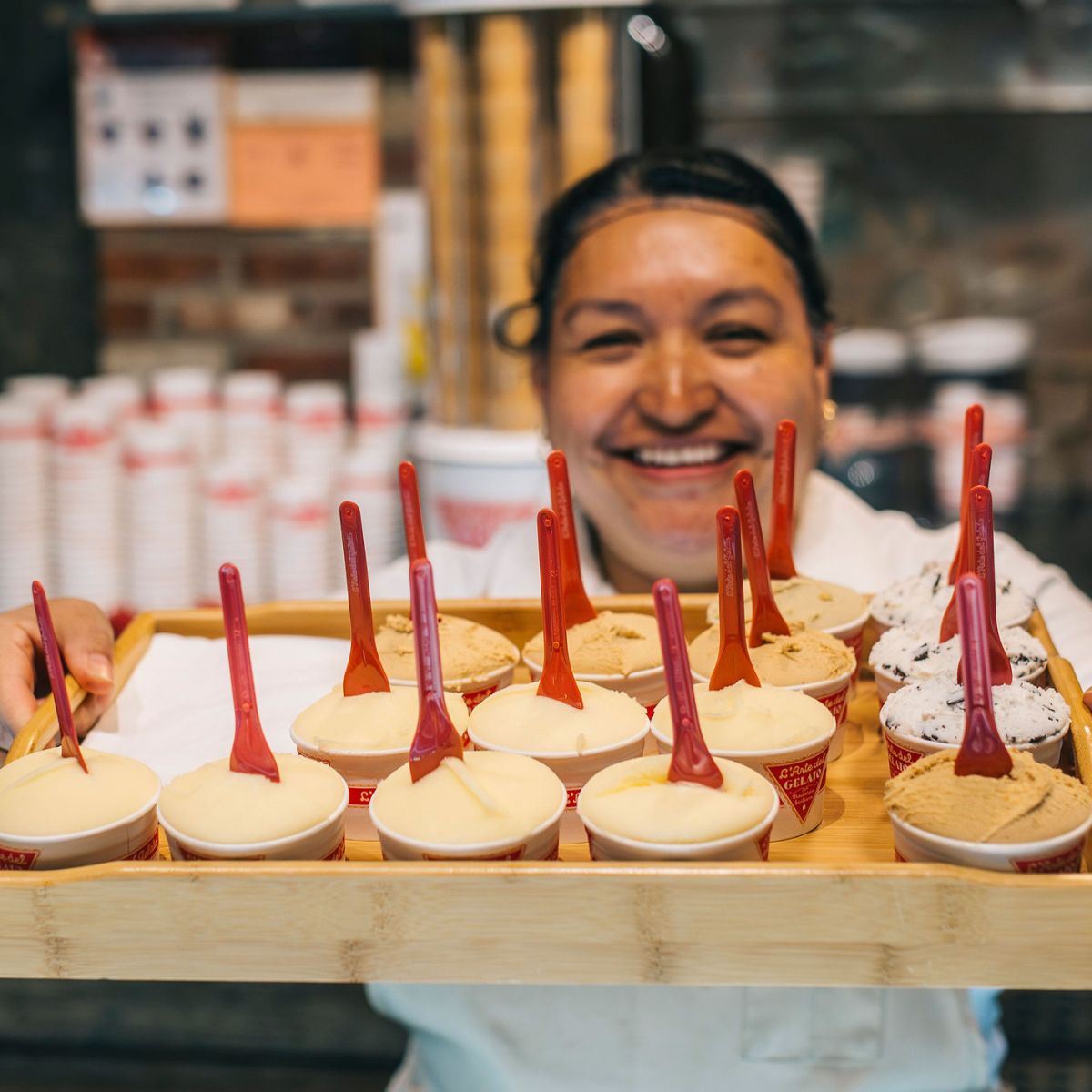Smiling person holding a tray of ice cream cups with red spoons.