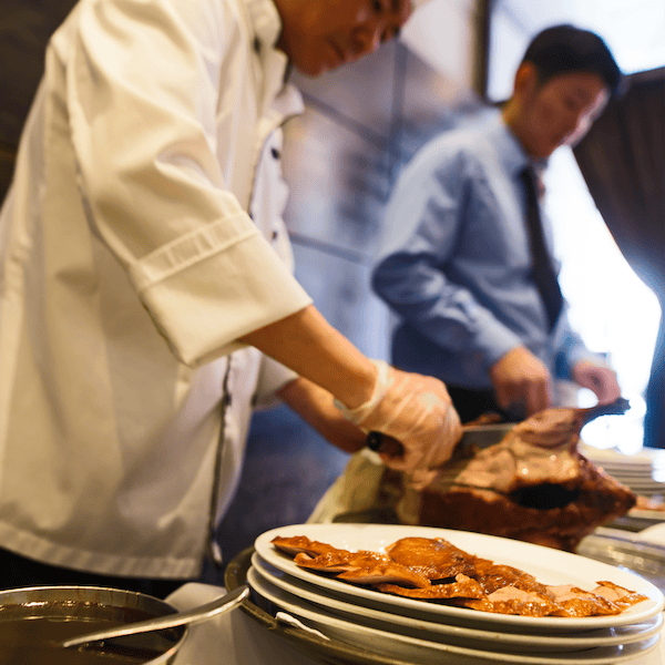Chef in white coat carving meat, plates of food in foreground, suited man in background.